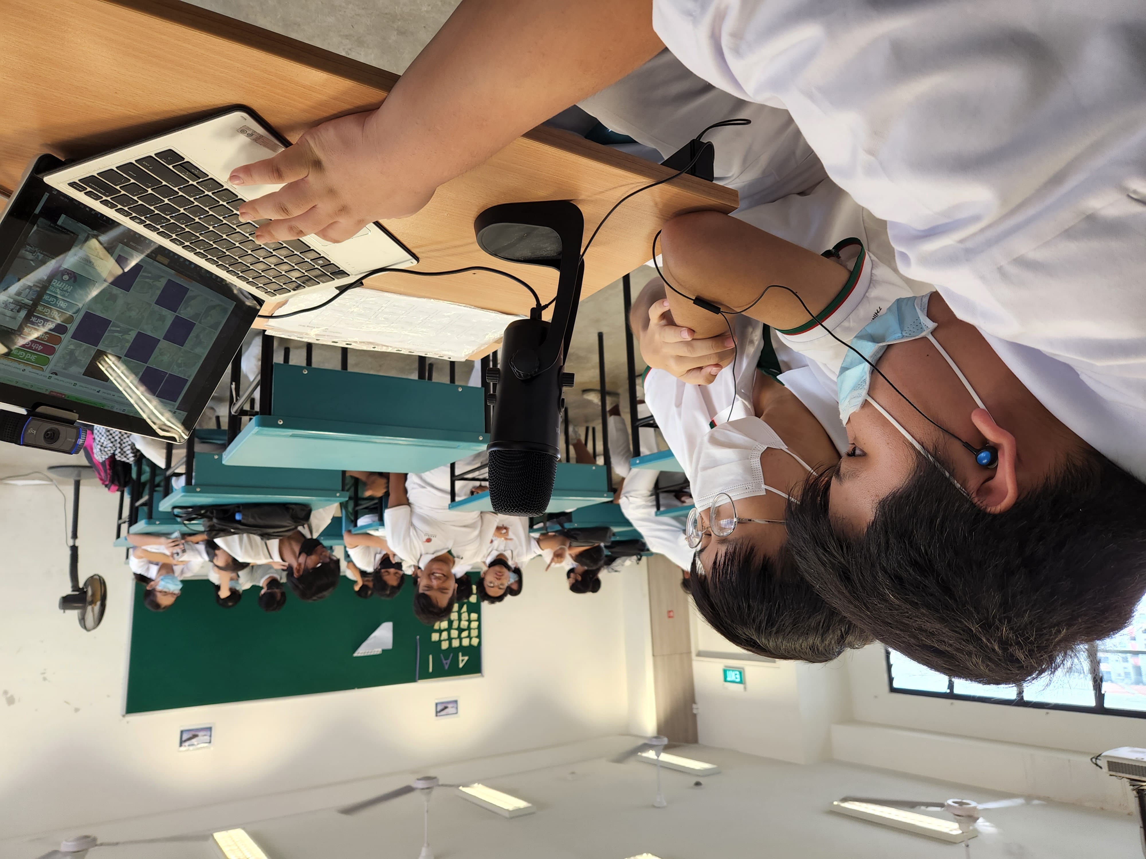 Two masked students use a laptop with a microphone at a desk in a classroom full of other students.